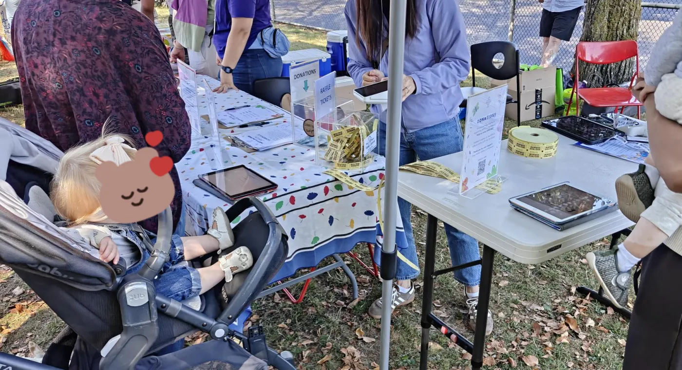A donation and raffle table at an outdoor event, with OpenCommunity tablets, a clear donations box, and rolls of raffle tickets.