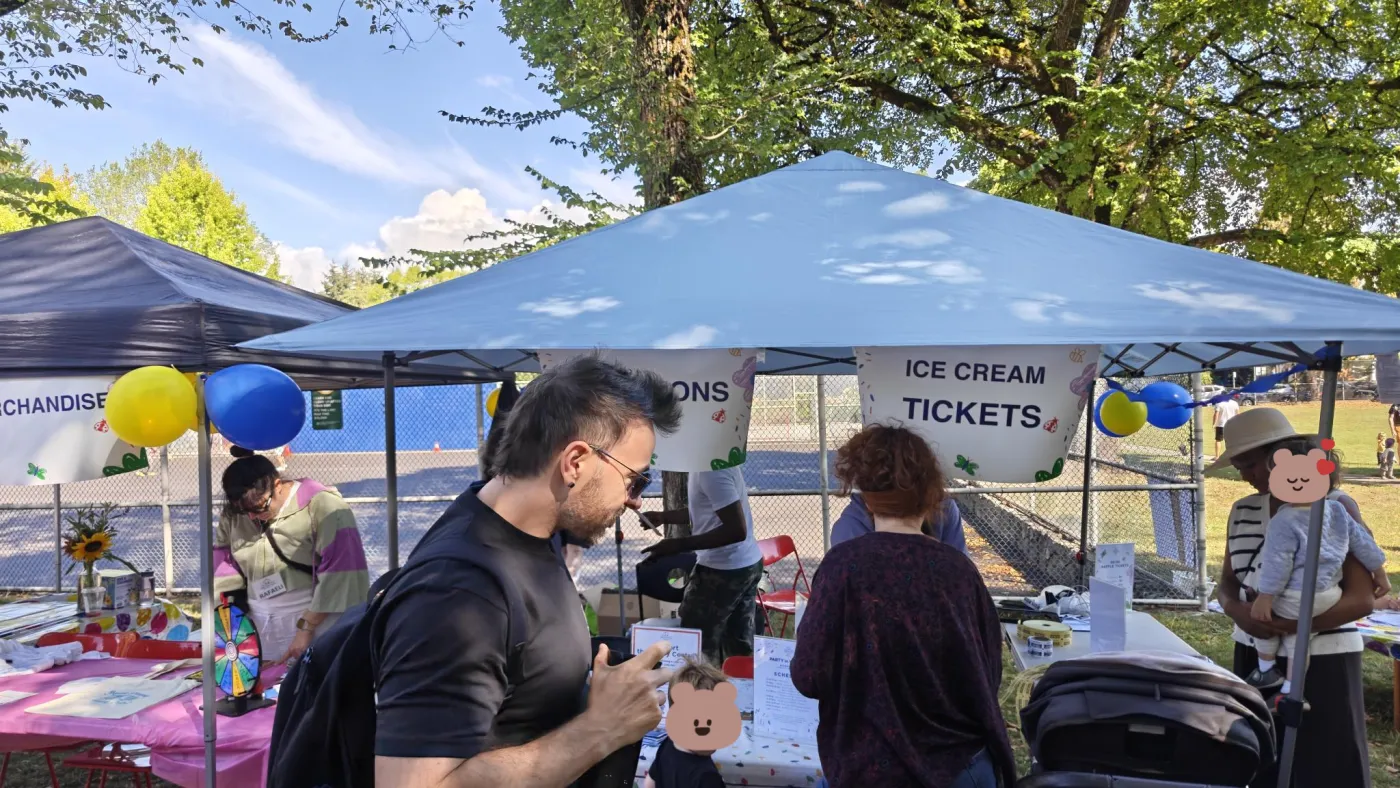 An outdoor event booth with Merchandise and Ice Cream Tickets banners, balloons, and families browsing in the sun.